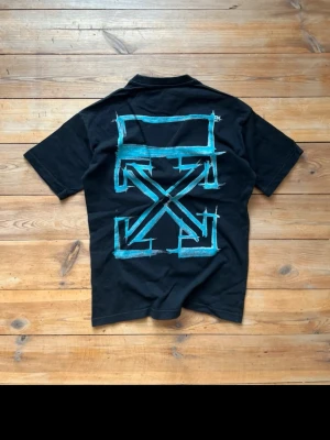 Svart Off-White t-shirt med blått tryck - Svart t-shirt från Off-White med stort blått grafiskt tryck på ryggen och liten text på bröstet. Klassisk passform med rund hals och korta ärmar. Tillverkad i mjuk bomull, perfekt för en streetwear-look. Priset är ej fast, buda gärna!
