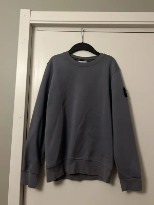 Grå sweatshirt från Stone Island - Säljer en stilren blå ish sweatshirt från Stone Island med klassisk rund halsringning och ribbade muddar. Tröjan har den ikoniska Stone Island-loggan på vänster ärm. Perfekt för dig som gillar streetwear och enkel design.