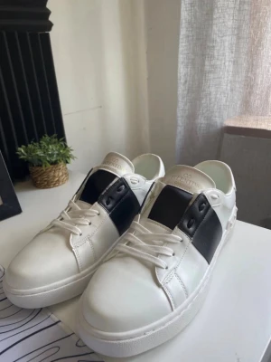 Vita och svarta sneakers från Valentino -  Helt oanvända bara testad inom hus vita sneakers från Valentino med svart panel på sidorna och klassisk snörning. Skorna har en stilren design med diskret logga på plösen och undersidan. Tillverkade i läder med gummisula för extra komfort.
