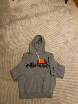 Grå Ellesse hoodie med logga - Snygg grå hoodie från Ellesse med stor logga framtill i svart, orange och rött. Klassisk känguruficka och huva med vit dragsko. Perfekt för en avslappnad streetstyle-look.