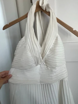 Vit långklänning med halterneck - Elegant vit långklänning med halterneck och djup v-ringning. Klänningen har plisserad överdel och kjol, vilket ger ett snyggt fall. Perfekt för sommarens speciella tillfällen. Tillverkad i ett lätt, luftigt material.