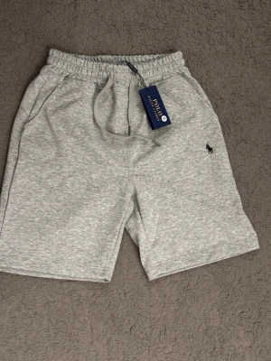 Ralph Lauren shorts - Snygga ljusgrå shorts från Polo Ralph Lauren med elastisk midja och snörning. Klassisk logga broderad på vänster ben och två sidofickor. Perfekta för chill dagar eller när du vill ha en clean och enkel stil.