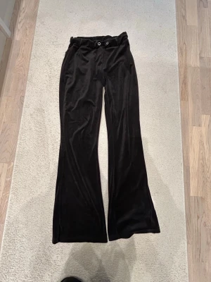 Svarta mjukisbyxor i velour - Säljer ett par svarta mjukisbyxor i mjuk velour med utsvängda ben och resår i midjan. Byxorna har dragsko med metallöljetter och en avslappnad passform. Perfekta för chill dagar eller när du vill vara extra bekväm.