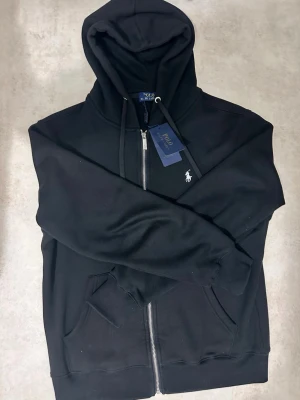 Svart hoodie från Polo Ralph Lauren - Snygg svart hoodie från Polo Ralph Lauren med dragkedja framtill, huva med dragsko och klassisk vit logga broderad på bröstet. Hoodien har kängurufickor och är tillverkad i mjukt bomullsmaterial som känns riktigt skönt mot huden.