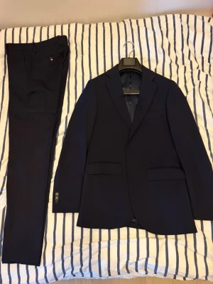 Mörkblå kostym från Riley - Snygg mörkblå kostym från Riley med klassisk kavaj och matchande byxor. Kavajen har två knappar, fickor med lock och slät finish. Perfekt student, avslutning, bal osv. Knappt använd.