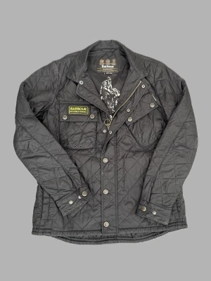 Svart quiltad jacka från Barbour - Snygg svart quiltad jacka från Barbour International med tryckknappar, dragkedja och två bröstfickor. Jackan har en klassisk krage, Barbour-märke på bröstet och är tillverkad i lätt, glansigt material. Perfekt för dig som gillar en tidlös och sportig look.