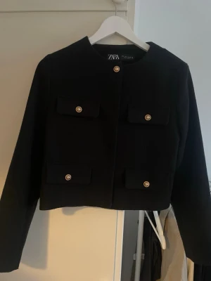 Svart cardiganjacka från Zara - Stilren svart cardiganjacka från Zara med rund halsringning och fyra dekorativa fickor framtill. Jackan har guldknappar som ger en elegant touch och är tillverkad i ett mjukt, vävt material. Perfekt för att lyfta vilken outfit som helst.
