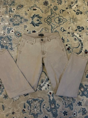 Beige chinos från LAGENCY - Snygga beige chinos från LAGENCY med klassisk passform och raka ben. Byxorna har bälteshällor, två snedställda fickor fram och knappstängning med LAGENCY-logga. Perfekta för en stilren och avslappnad look.