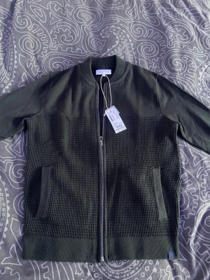 Mörkgrön cardiganjacka Tom Tailor - Snygg mörkgrön cardiganjacka från Tom Tailor med ribbad krage och hel dragkedja. Jackan har två fickor framtill och ett coolt stickat rutmönster på nedre delen. Perfekt för att ge din outfit en avslappnad men stilren touch.