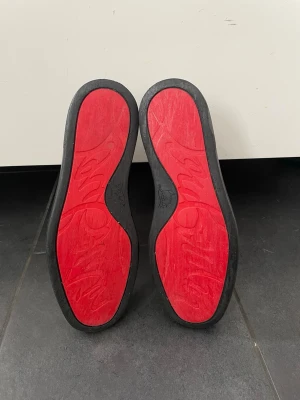 Svarta Christian Louboutin skor - Snygga svarta sneakers från Christian Louboutin med klassisk röd sula. Skorna har snörning och är tillverkade i mocka med svarta detaljer. Perfekt för dig som vill ha en stilren och lyxig look.