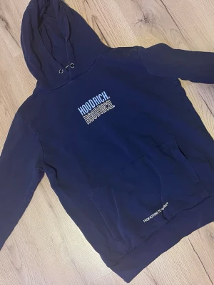 Mörkblå hoodie från Hoodrich - Mörkblå hoodie från Hoodrich med tryck på både bröstet och ryggen. Stor känguruficka framtill och texten 'FROM NOTHING TO SOMETHING' tryckt nedtill. Klassisk huva med dragsko och avslappnad passform. Perfekt för en streetwear-look.