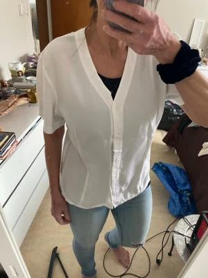 Blus  från NO 1   med v-ringning - Snygg vit blus med v-ringning och korta ärmar. Blusen har knäppning framtill och en avslappnad passform som passar perfekt till jeans eller kjol. Tillverkad i ett lätt och luftigt material som känns skönt mot huden.