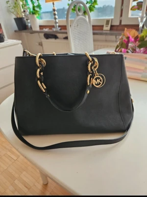 Svart ÄKTA Michael Kors handväska - Snygg svart handväska från Michael Kors med guldfärgade detaljer och kedjor. Väskan har både handtag och en längre axelrem, samt en rund MK-logga i metall. Tillverkad i slätt läder och har en klassisk, rymlig form. Kan finnas små spår av defekter men syns knappt😊Äkta