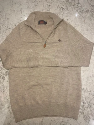 Beige merinoulltröja från Morris - Snygg beige stickad tröja från Morris i 100% merinoull. Tröjan har halv dragkedja, hög krage och diskret Morris-logga broderad på bröstet. Perfekt för dig som gillar stilrena och klassiska plagg med en modern touch.