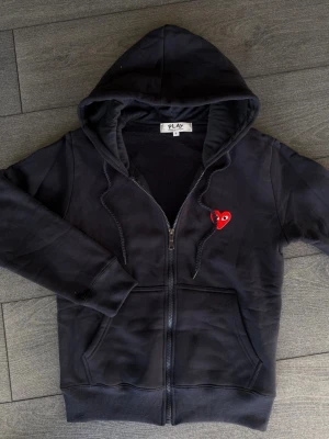 Marinblå  hoodie från Comme des Garçons Play - Snygg svart hoodie med dragkedja från Comme des Garçons Play. Klassisk huva med dragsko och två fickor framtill. Ikoniskt rött hjärta med ögon broderat på bröstet. Tillverkad i mjuk bomull för skön komfort.