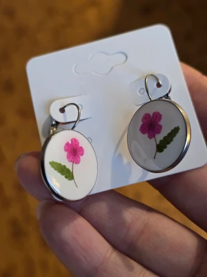 Runda örhängen med rosa blomma - Unika runda örhängen med infattade rosa blommor och gröna blad på vit bakgrund. Perfekta för dig som gillar naturinspirerade accessoarer och vill ha något som sticker ut. Rostfritt stål.