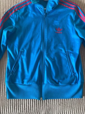 Gammal adidas tröja - Säljer en blå ziptröja från Adidas med klassiska rosa ränder längs ärmarna och rosa broderad logga på bröstet. Tröjan har ribbade muddar, dragkedja framtill och två fickor. Perfekt för en sportig och retro look.
