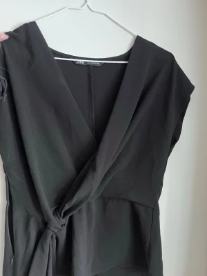 Svart omlottopp från Zara - Stilren svart omlottopp från Zara med v-ringad front och knyt i midjan. Toppen har korta ärmar och en elegant, figurnära passform. Perfekt för dig som gillar minimalistisk och trendig stil. Storlek S