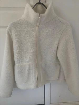 Vit teddyjacka med dragkedja - Mysig vit teddyjacka med hög krage och hel dragkedja framtill. Jackan har två stora fickor på framsidan och är gjord i fluffigt, mjukt teddy-material. Perfekt för att hålla värmen under kyliga dagar och ger en trendig look.