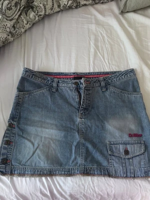 Kort jeanskjol från Dobber  - Cool kort jeanskjol från Dobber med ficka och knappar på sidan. Kjolen har broderade detaljer bak och bälteshällor. Perfekt för dig som gillar en avslappnad och trendig look. Den klassiska blå denimfärgen gör den enkel att matcha. Storleken säger 32 men passar en M
