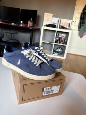 Blåa Polo Ralph Lauren sneakers - Snygga blåa sneakers från Polo Ralph Lauren med vit logga på sidan och vita snören. Skorna har en låg profil och ovandel i mocka, samt en beige sula med Polo-tryck. Perfekta för en clean och stilren look.