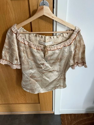 Beige silkesblus från Munthe & Simonsen - Romantisk beige Silkesblus från Munthe & Simonsen. Toppen har korta puffärmar, snörning framtill och volangdetaljer längs urringningen. Mönstrad med subtila blommor och skimrande yta. Perfekt för dig som gillar unika detaljer.