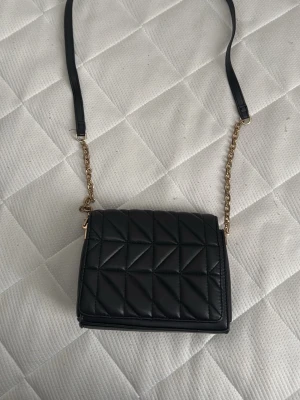 Svart quiltad axelväska med kedja - Snygg svart axelväska i quiltat mönster med guldfärgad kedja och justerbar axelrem i konstläder. Väskan har en stilren, rektangulär form och stängs med lock. Perfekt för dig som vill ha en trendig och praktisk accessoar.