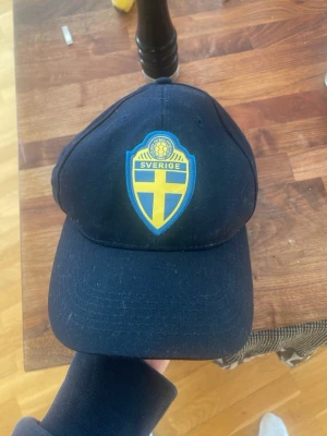 Mörkblå Sverige keps med brodyr - Snygg mörkblå keps med broderad Sverige-logga framtill och texten 'SVERIGE' i gult baktill. Klassisk böjd skärm och justerbar rem bak. Perfekt för att visa ditt stöd för Sverige, sportig och stilren design.