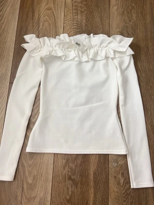 Vit volangtop från NLY Trend - Snygg vit topp från NLY Trend med långa ärmar och bred volang runt axlarna. Offshoulder-modell som ger en trendig look. Perfekt att styla med jeans eller kjol för en chic outfit.