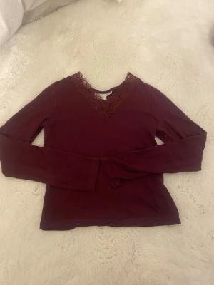 Vinröd långärmad topp med spets - Vinröd långärmad topp med v-ringning och dekorativ spets runt halsen. Toppens material känns mjukt och stretchigt, perfekt för en snygg och bekväm look. Passar dig som gillar stilrena detaljer och en mörkare färgton.