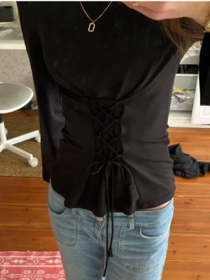 Svart topp med snörning i midjan - Snygg svart topp med snörning framtill i midjan som ger en korsettkänsla. Modellen är figurnära och har långa ärmar. Perfekt att styla med jeans eller kjol för en trendig look.