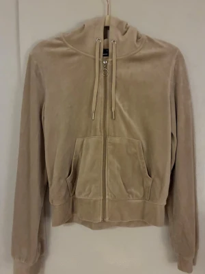 Beige hoodie med dragkedja - Mjuk beige hoodie med dragkedja framtill, fickor och huva med snören. Långärmad modell med ribbade muddar och avslappnad passform. Perfekt för en chill och bekväm stil.