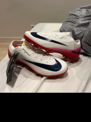 Nike Mercurial vita elite fotbollsskor - Snygga Nike Mercurial fotbollsskor i vitt med stora svarta swoosh-loggor på sidorna och rosa/röd sula. Skorna har låg profil, snörning och är tillverkade i ett lätt syntetmaterial för bästa känsla och grepp på planen. Perfekta för dig som vill sticka ut på matchen.