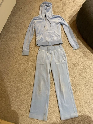 Juicy Couture Ljusblå velour loungewear-set - Mysigt loungewear-set i ljusblå velour med huva och dragkedja på tröjan samt breda byxben och dragsko i midjan. Perfekt för chill dagar hemma. Setet har stora fickor på byxorna och ribbade muddar på tröjan. Byxorna är upplagda och passar upp till ca 1,55 m. Tröjan är som ny byxorna fina men mer använt skick.