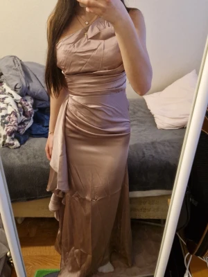 Champagnefärgad långklänning oneshoulder - Elegant långklänning i champagnefärg med oneshoulder-design och draperade detaljer. Klänningen har en figurnära passform och ett vackert fallande tyg med volangdetalj på sidan. Perfekt för speciella tillfällen.