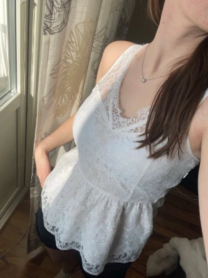 Vit spetsblus med peplum - Superfin vit blus i spets med peplumskärning och ärmlös design. Snygg v-ringning med extra spetsdetaljer upptill. Perfekt att styla med jeans eller kjol för en fräsch look.