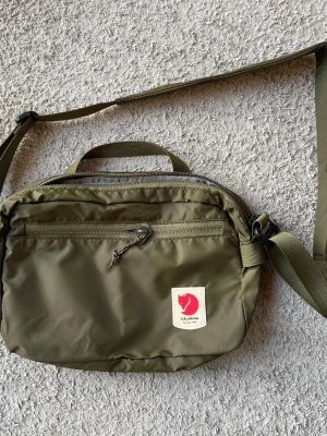 Fjällräven High Coast Crossbody - Jättefin grön axelremsväska från Fjällräven.   I nyskick.   Text från Naturkompaniet:  Produktbeskrivning Lätt och liten axelväska Lätt och smidig axelväska för resor, snabba ärenden på stan eller som komplement till en ryggsäck. Den vadderade axelremmen går att ta av och väskan kan därmed också användas som exempelvis necessär.  High Coast Crossbody rymmer 3 liter och har två fickor med dragkedja, en invändig och en utvändig. Inuti fins även två meshfack för ytterligare organisering av dina vär