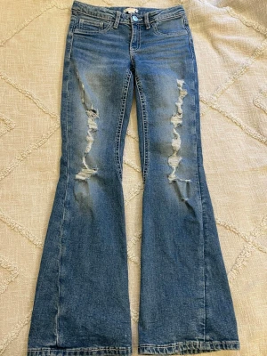 Blå bootcut jeans med slitningar - Säljer ett par blå bootcut jeans från Gina Young med coola slitningar framtill och snygga detaljerade bakfickor med knappar. Jeansen har medelhög midja. Perfekta för dig som gillar en avslappnad och trendig stil. Storlek 164