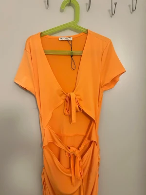 Orange omlottklänning från NLY One - Säljer en orange kortklänning från NLY One med omlottdesign, knytband i midjan och vid bysten. Klänningen har korta ärmar, djup v-ringning och volangdetalj nedtill. Perfekt för sommarens alla tillfällen!