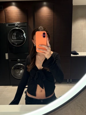 Svart croppad omlottopp med knyt - Trendig svart croppad topp med omlott-design och knyt framtill. Långärmad och djup v-ringning, perfekt för att skapa en cool och stilren look. Passar dig som gillar att visa lite midja och ha en edgy stil.