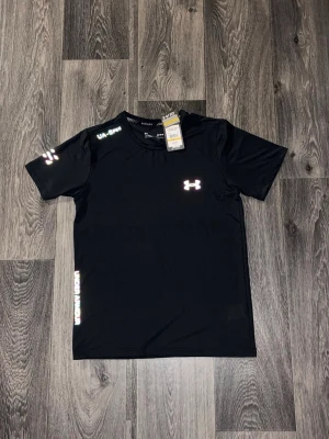 Svart tränings-tshirt från Under Armour - Snygg svart tränings-tshirt från Under Armour med reflekterande logga på bröstet och detaljer på ärmen och axeln. Tillverkad i lätt och snabbtorkande polyester, perfekt för intensiva träningspass. Klassisk passform och korta ärmar.