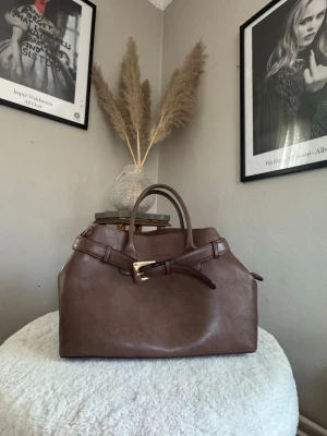 Brun handväska med gulddetalj - Stilren brun handväska i slätt läder med två handtag och dekorativt bälte med guldfärgat spänne framtill. Väskan har en klassisk, något boxig form och passar perfekt för dig som gillar tidlös design och snygga detaljer.