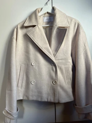 Beige kort rock från Bubbleroom - Stilren och kort beige rock från Bubbleroom med dubbelknäppning och bred krage. Rocken har raka fickor och stora knappar framtill. Perfekt för dig som gillar en klassisk och trendig look. Materialet är mjukt och har en lätt ullkänsla.