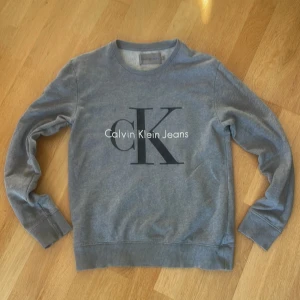 Grå sweatshirt från Calvin Klein Jeans - Snygg grå sweatshirt från Calvin Klein Jeans med stort CK-tryck på bröstet. Tröjan har rund hals, långa ärmar och ribbade muddar vid ärmslut och nederkant. Perfekt för en chill och stilren look.