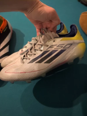 Adidas F50 Elite stl 39 1/3del - Säljer ett par Adidas F50 Elite fotbollsskor i vitt med svarta och blå detaljer samt gula inslag vid hälen. Storlek 39 1/3del. Bra skick men välanvända, dobbar i bra skick se bilder