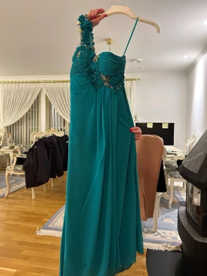Turkos långklänning med blommiga detaljer - Elegant turkos långklänning med enaxlad design och smal axelrem. Klänningen har vackra blommiga applikationer och glittrande stenar längs axeln och bysten. Tillverkad i ett lätt, flytande material som ger ett lyxigt intryck. Perfekt för speciella tillfällen.