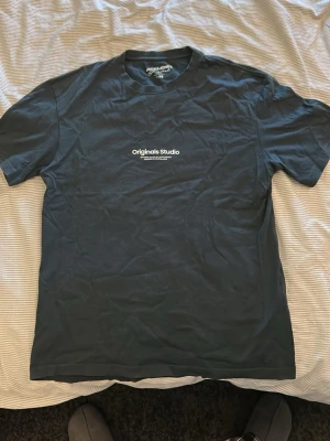 Mörkblå t-shirt från Jack & Jones - Snygg mörkblå t-shirt från Jack & Jones Originals Studio med vit text på bröstet. Klassisk passform och rund halsringning. Perfekt till jeans eller shorts för en clean och enkel stil.