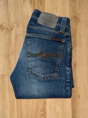 Nudie jeans  - Säljer ett par blå nudie jeans i modellen Tight long john. Jeansen har en mörk tvätt med lätt slitning. Midja 31cm, längd 97cm. JAG SKICKAR EJ FLER BILDER NÄR JAG BÄR JEANSEN! för bättre inblick i passformen rekommenderar jag att googla på modellnamnet. För storleksguide kolla måtten noggrant då jeansen alltid kan vara uppsydda heller krympta och jag ej tar emot returer :) s5,3