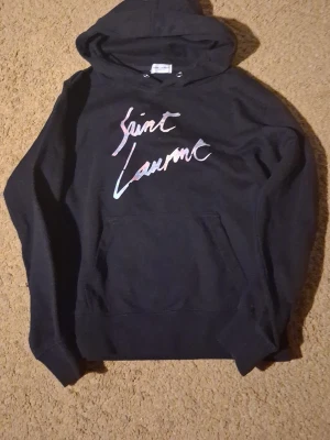 Svart Saint Laurent hoodie - Snygg svart hoodie från Saint Laurent med coolt färgat tryck i rosa och blått på bröstet. Klassisk känguruficka framtill och huva med dragsko. Perfekt för dig som vill ha en stilren men ändå unik look.