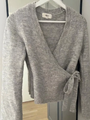 Grå kofta Gina tricot  - Mysig grå omlottkofta med knyt i sidan och långa ärmar. Tröjan har en ribbad kant nedtill och vid ärmsluten, vilket ger en snygg och enkel look. 